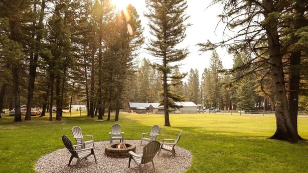 A circular seating area with chairs around a fire pit, surrounded by a gravel circle on a grassy field, with tall trees and a building in the background. - Convenient Mountain Comfort (Lincoln)