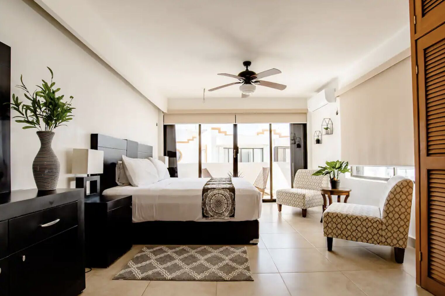 A modern bedroom with a bed, bedside tables, a ceiling fan, a chair, a small table, and a potted plant.