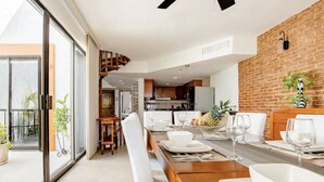Un espace repas moderne avec une table en bois dressée pour quatre personnes, une cuisine en arrière-plan et un mur de briques agrémenté d’une plante.