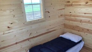 A small, single-bed room with a wooden floor and walls, a window, and a simple bed with a dark blanket and white pillow. - Valley Cabin (Spring Valley)