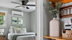 Un dormitorio con cama, ventilador de techo, mesita de noche y tocador. Se ve una ventana con persianas, un aire acondicionado de pared y un estante con toallas y cestas.