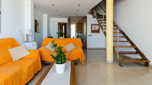 A modern living room with two orange sofas, a white coffee table with a plant, a wooden staircase, and a kitchen in the background. - YourHouse Mulet, cozy sea-view apartment (Can Picafort)