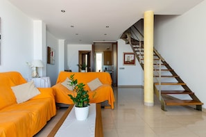 Ein modernes Wohnzimmer mit zwei orangenen Sofas, einem weißen Couchtisch mit Pflanze, einer Holztreppe und einer Küche im Hintergrund.