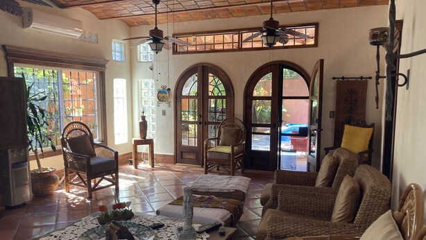 A spacious living area with a high ceiling, multiple seating options including a sofa and chairs, a coffee table with decorative items, and a view through large arched windows. - Casa Fiesta, Best Place to Rest, 1 Block Away From the Beach Owner Managed (Rincón de Guayabitos)