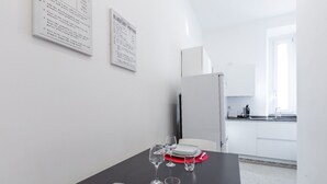 Uma área de refeições com uma mesa posta para dois, uma cozinha ao fundo e uma parede branca com um cardápio emoldurado.