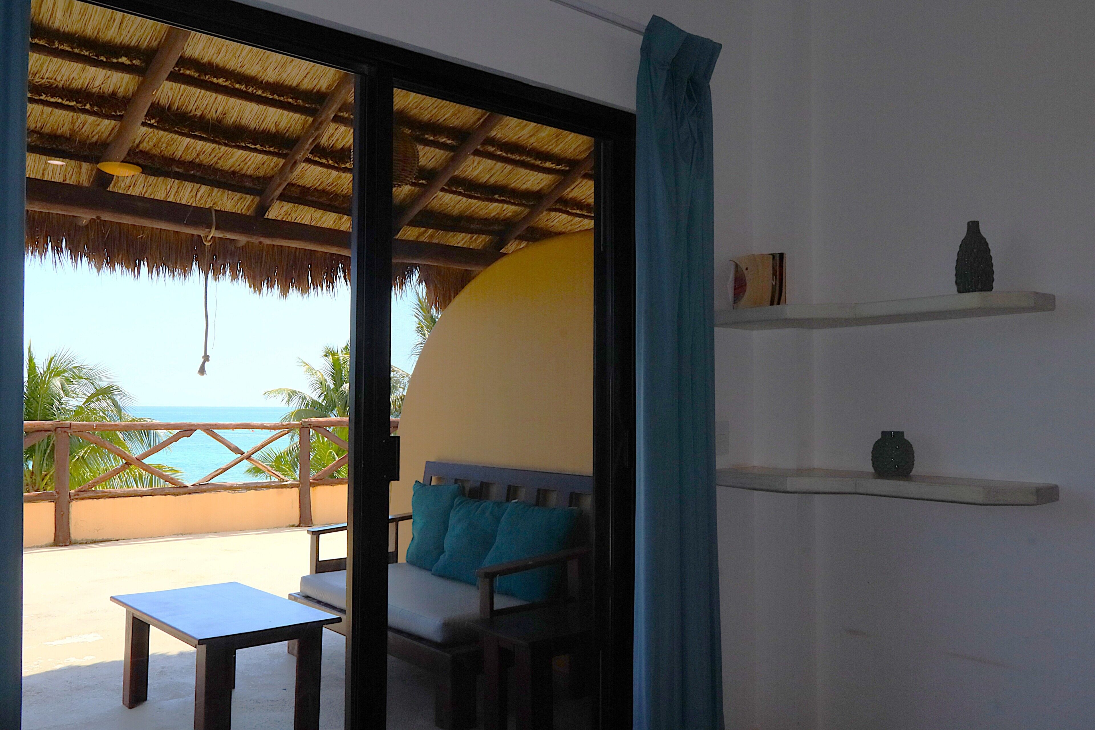 A room with a view of the sea, a thatched roof, a sofa with cushions, a table, and shelves with decorative items.