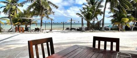 A wooden table with a laptop and a glass of wine, overlooking a beach with palm trees and the ocean.