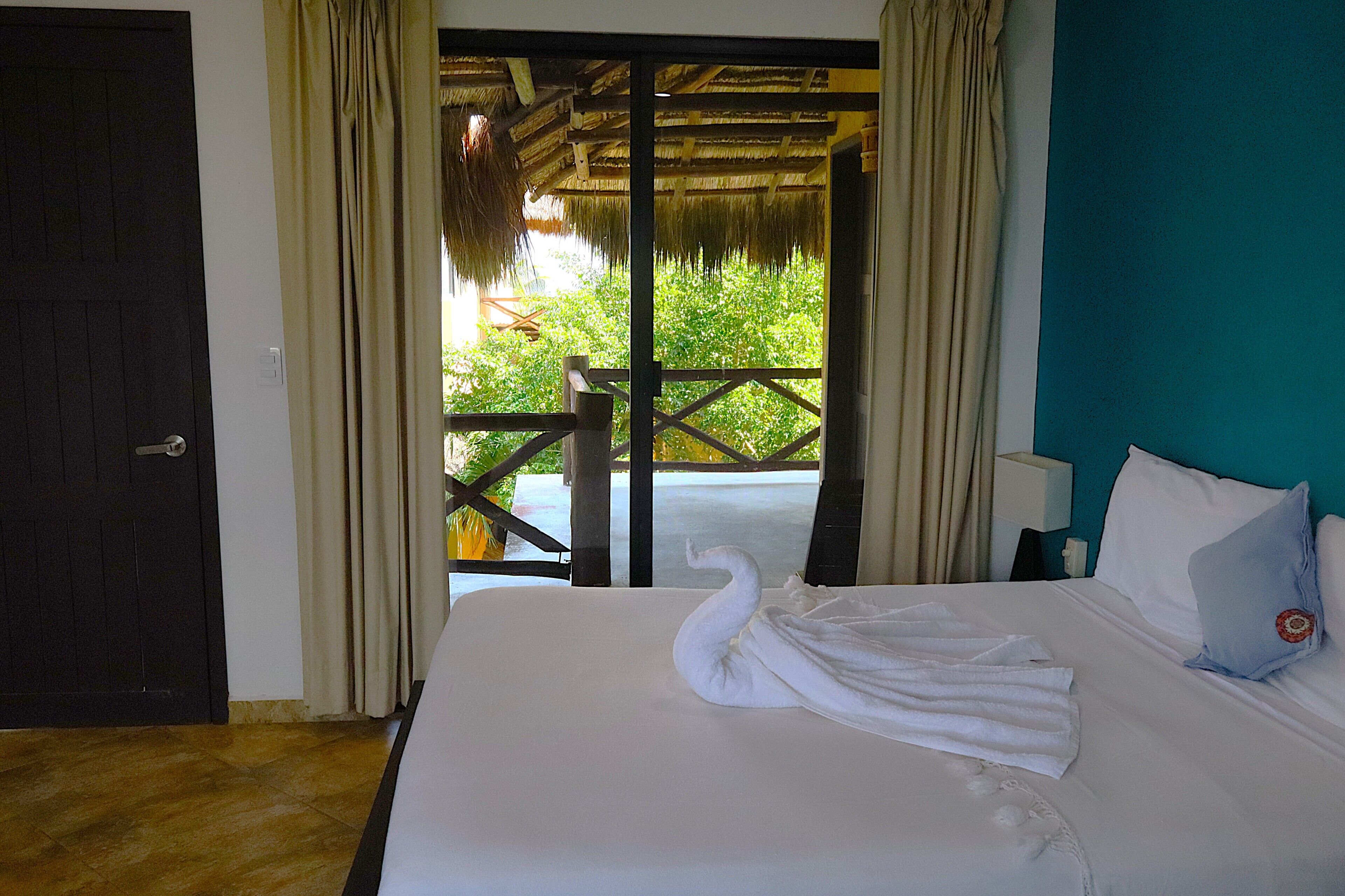 A hotel room with a bed, a swan-shaped towel, a teal wall, and a view of greenery through a door.