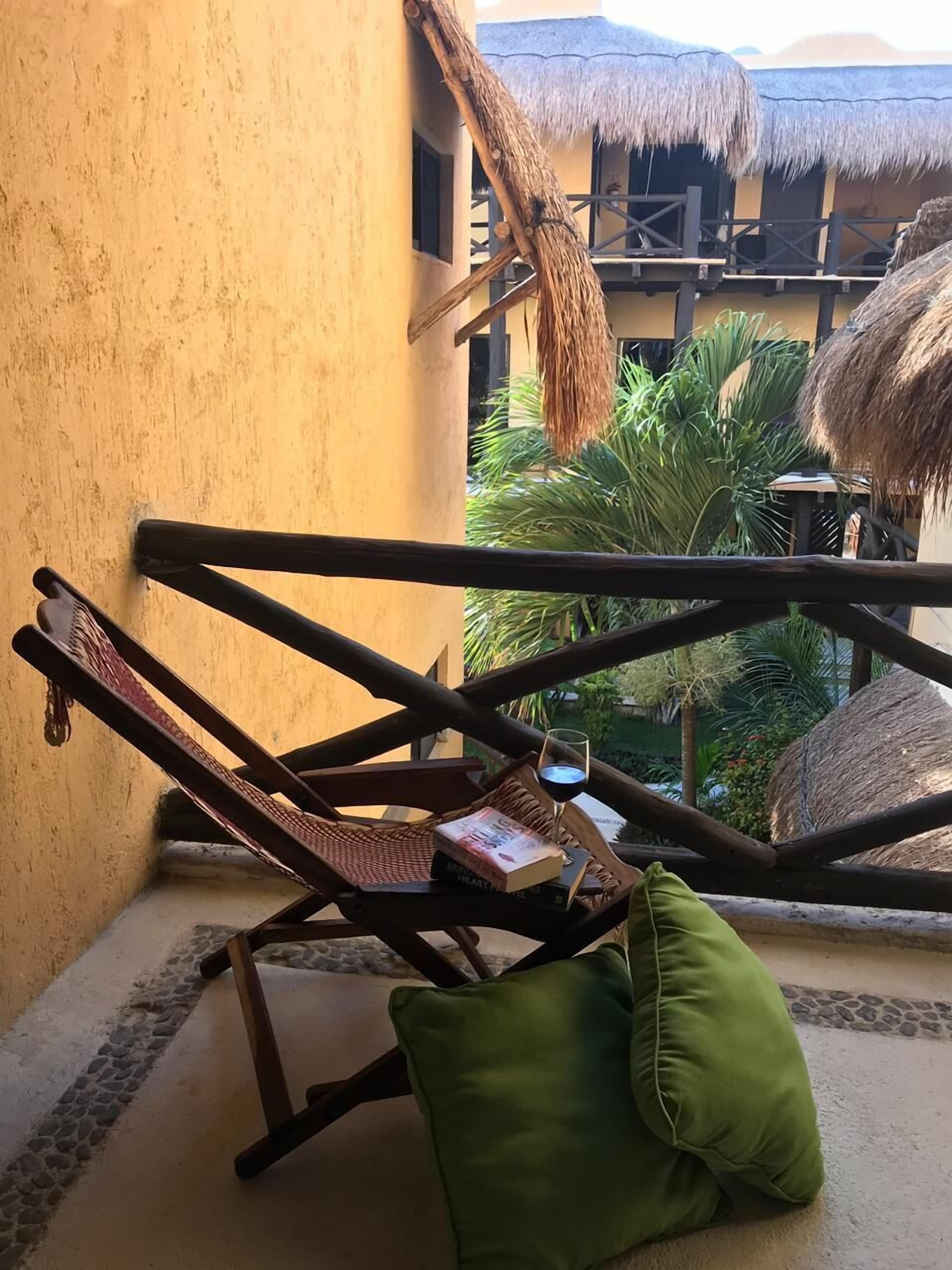 A wooden lounge chair with a green cushion, a book, and a glass of red wine on a balcony with a thatched roof and a view of greenery.
