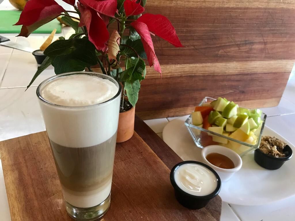 A glass of a layered beverage, a small bowl of sauce, and a fruit dish on a wooden table with a potted plant in the background.