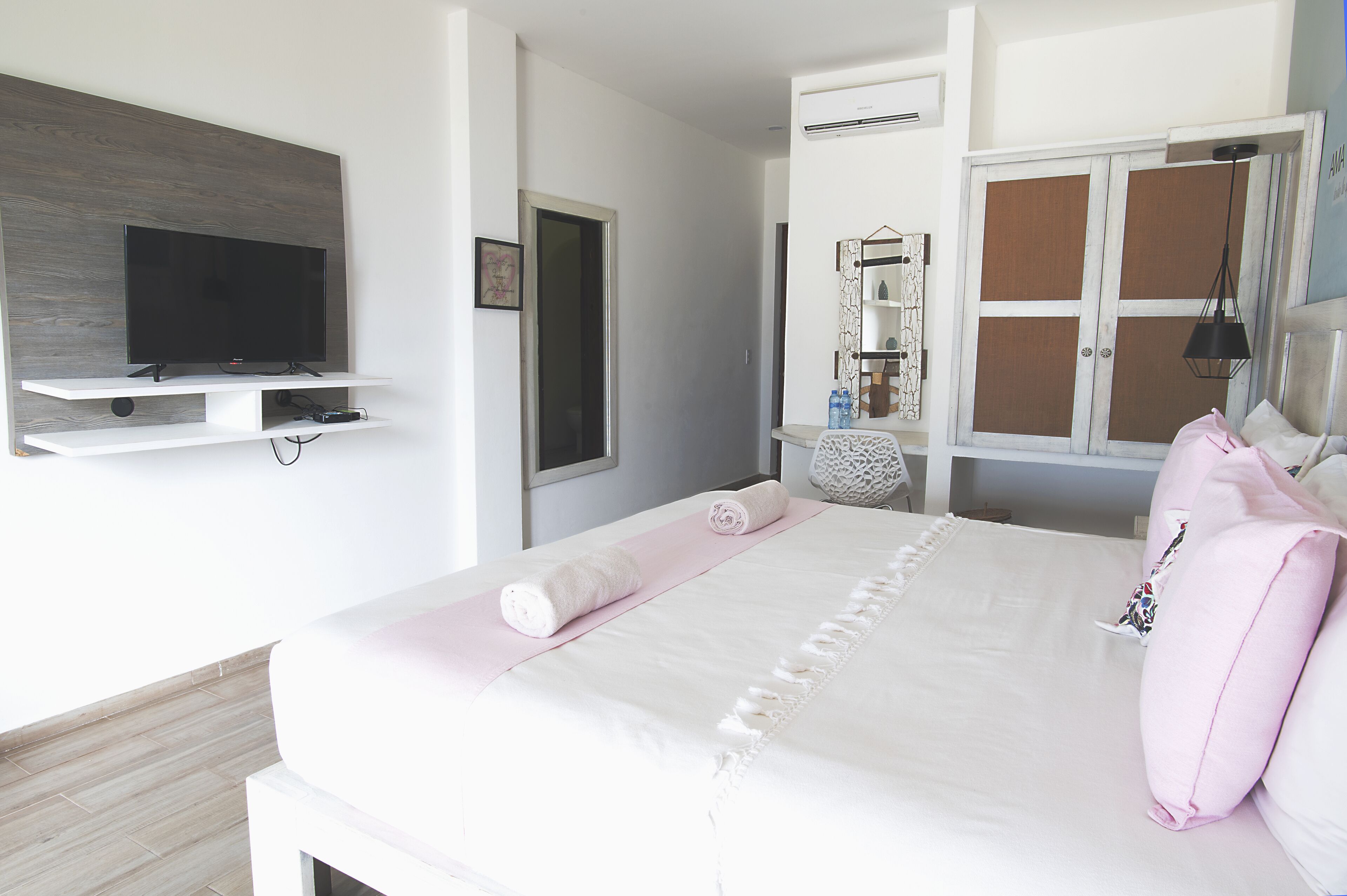A neatly arranged bedroom with a flat-screen TV mounted on a wall, a bed with a pink towel and pillows, a bedside table, and a wardrobe with a mirror.