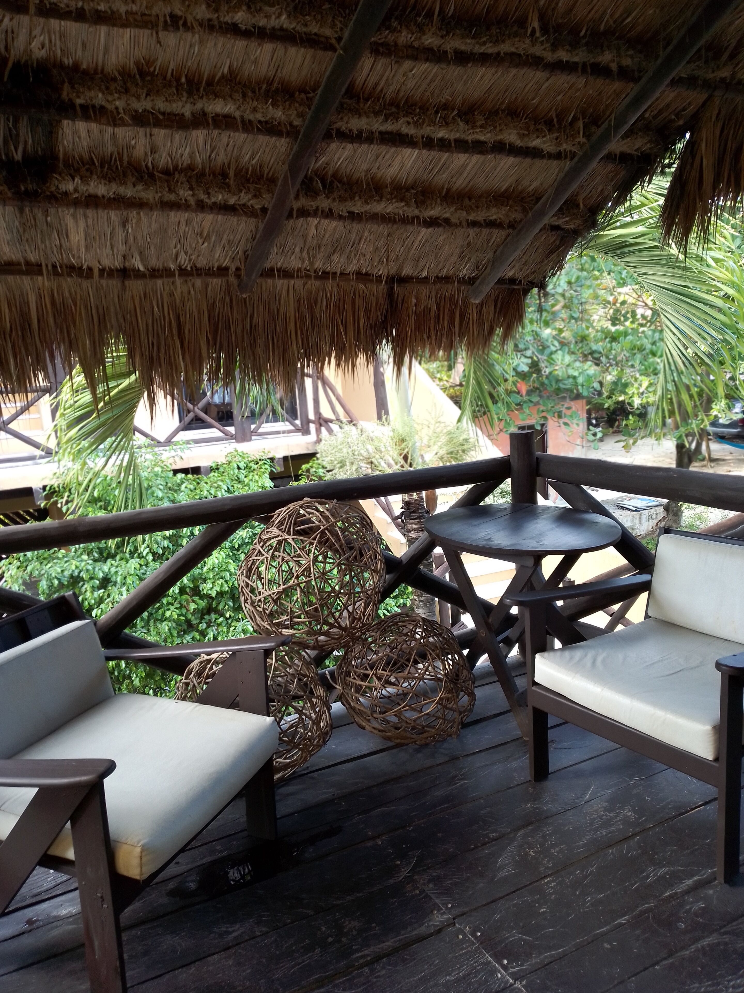 An outdoor seating area with wicker chairs and round tables, a thatched roof, and lush greenery in the background.