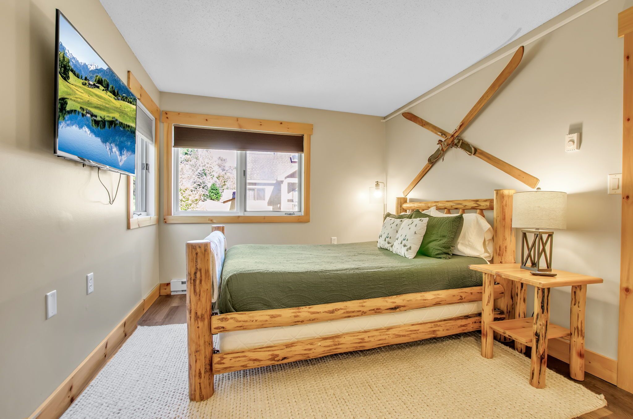 A bedroom with a wooden bed, a window with a view, a mounted TV, and a wooden ceiling beam.