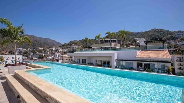 Una piscina en la azotea con agua azul cristalina, rodeada de edificios modernos y palmeras, con vistas a un paisaje montañoso.