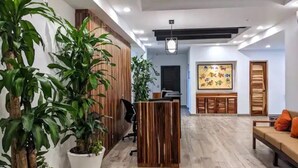 Un área de recepción con un escritorio de madera, plantas en macetas, un banco y una obra de arte de colores en la pared.