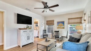 A living room with a television, a ceiling fan, a dining area with chairs, and a sofa with a blue pillow.