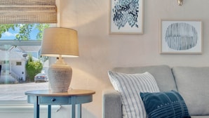 A living room with a lamp on a side table, a sofa with patterned cushions, and two framed artworks on the wall.