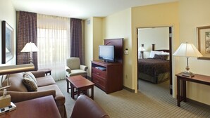 A hotel room with a sofa, armchair, TV, desk, lamp, and a bed with a headboard.