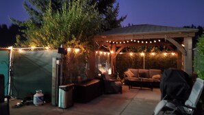 An outdoor patio area with a covered seating area, string lights, and a large tree. - Main floor studio well-furnished room.  Unit 2 with bath/shower bidet toilet (Pinehurst)