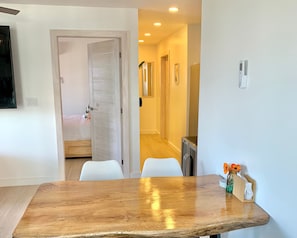 Una habitación de hotel moderna con una mesa de comedor de madera, paredes blancas y un pasillo que conduce a un baño.