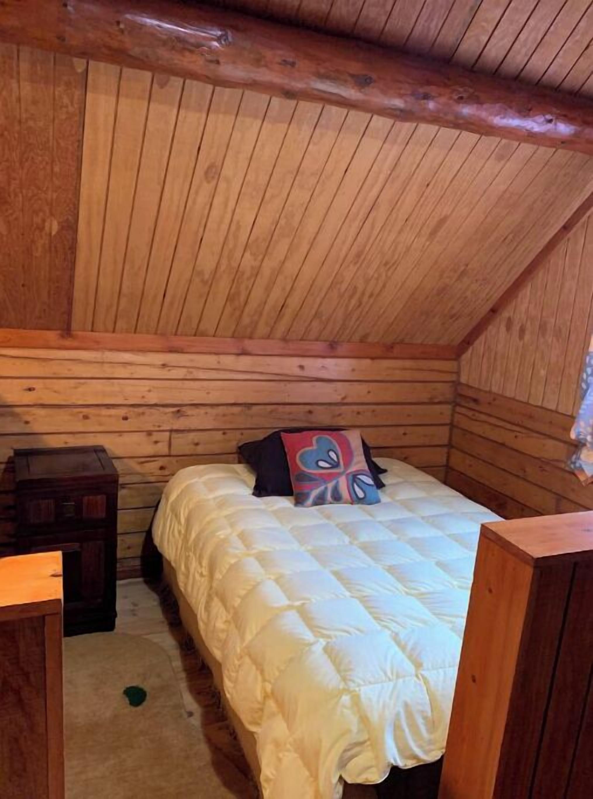 Une petite pièce chaleureuse aux murs en bois et au plafond en pente. On y trouve un lit simple avec une couette blanche et un coussin coloré, une petite table de chevet en bois et un banc en bois.