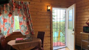 A wooden cabin interior with a dining table, chairs, a small TV on a stand, and a door leading to a balcony with a view of greenery. - Cabin 1 on the Kenai River!best view cabin (Sterling)