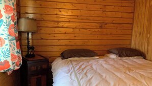 A wooden cabin room with a bed, a nightstand with a lamp, and a floral curtain. - Cabin 1 on the Kenai River!best view cabin (Sterling)