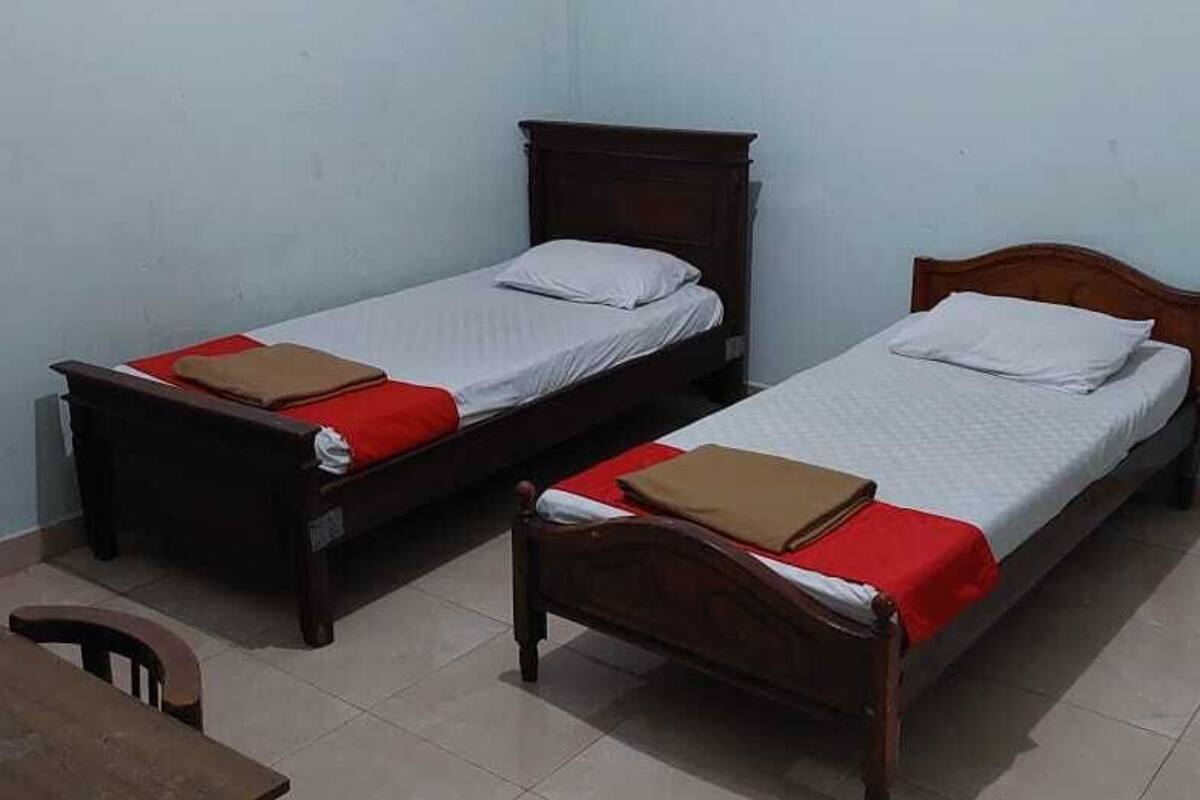 A room with two single beds, each with a wooden frame and red bedding. There is a small table and a chair in the corner.