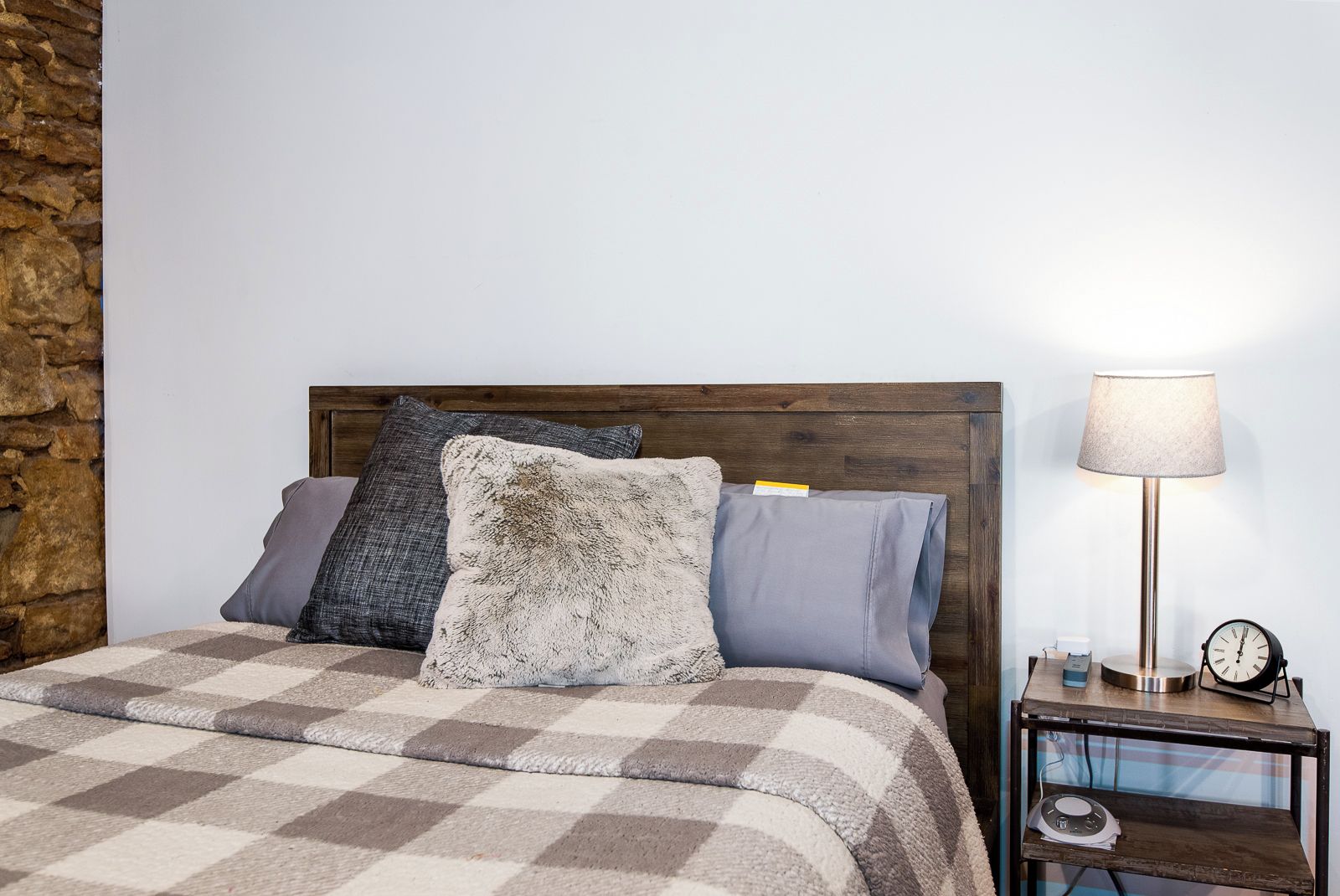 Una cama con una manta a cuadros, cabecera de madera y tres almohadas. Junto a la cama, una mesita de noche con una lámpara y un reloj.
