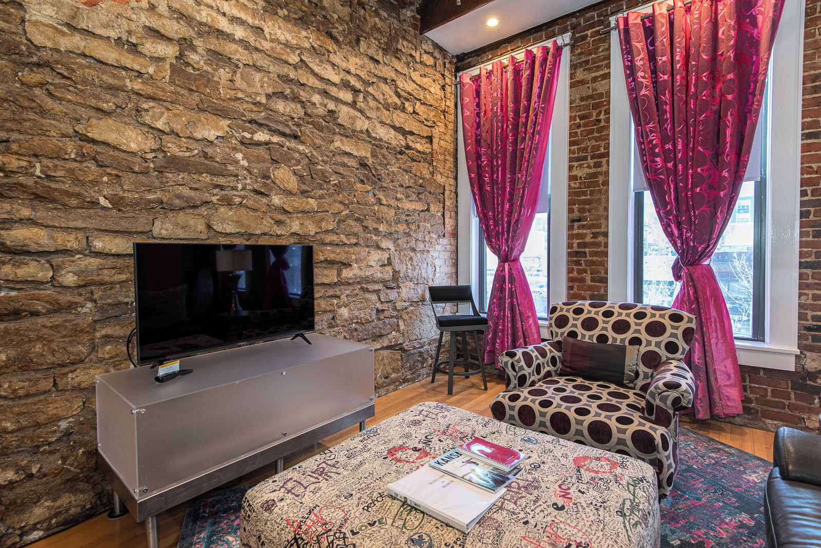 Un salon chaleureux avec un mur en pierre, un téléviseur à écran plat, un canapé à motifs et une fenêtre ornée de rideaux rouges.