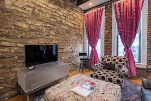 A cozy living room with a stone wall, a flat-screen TV, a patterned sofa, and a window with red curtains. - Suite Life en Mass #1 License #STR-20-00014 (Lawrence)