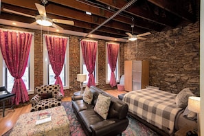 A cozy bedroom with a brick wall, a bed with a checkered blanket, a leather sofa, and a patterned ottoman.