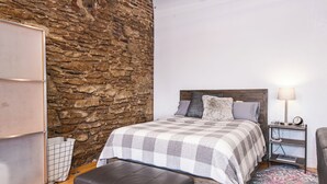 Un dormitorio con pared de piedra, una cama con manta a cuadros, una mesita de noche con lámpara y una silla.