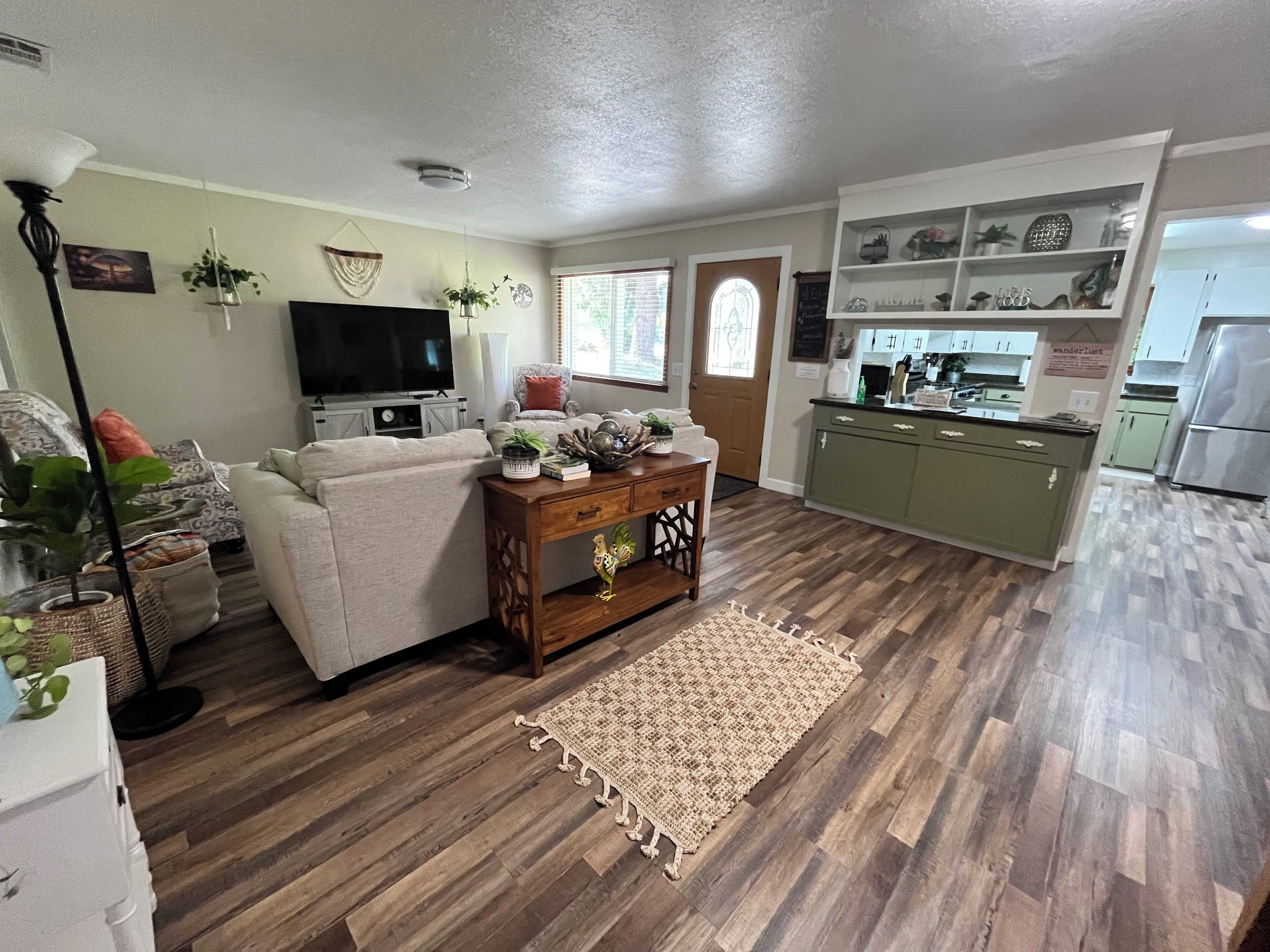 Una sala de estar con sofá, televisión y elementos decorativos, una cocina con armarios verdes y suelo de madera.