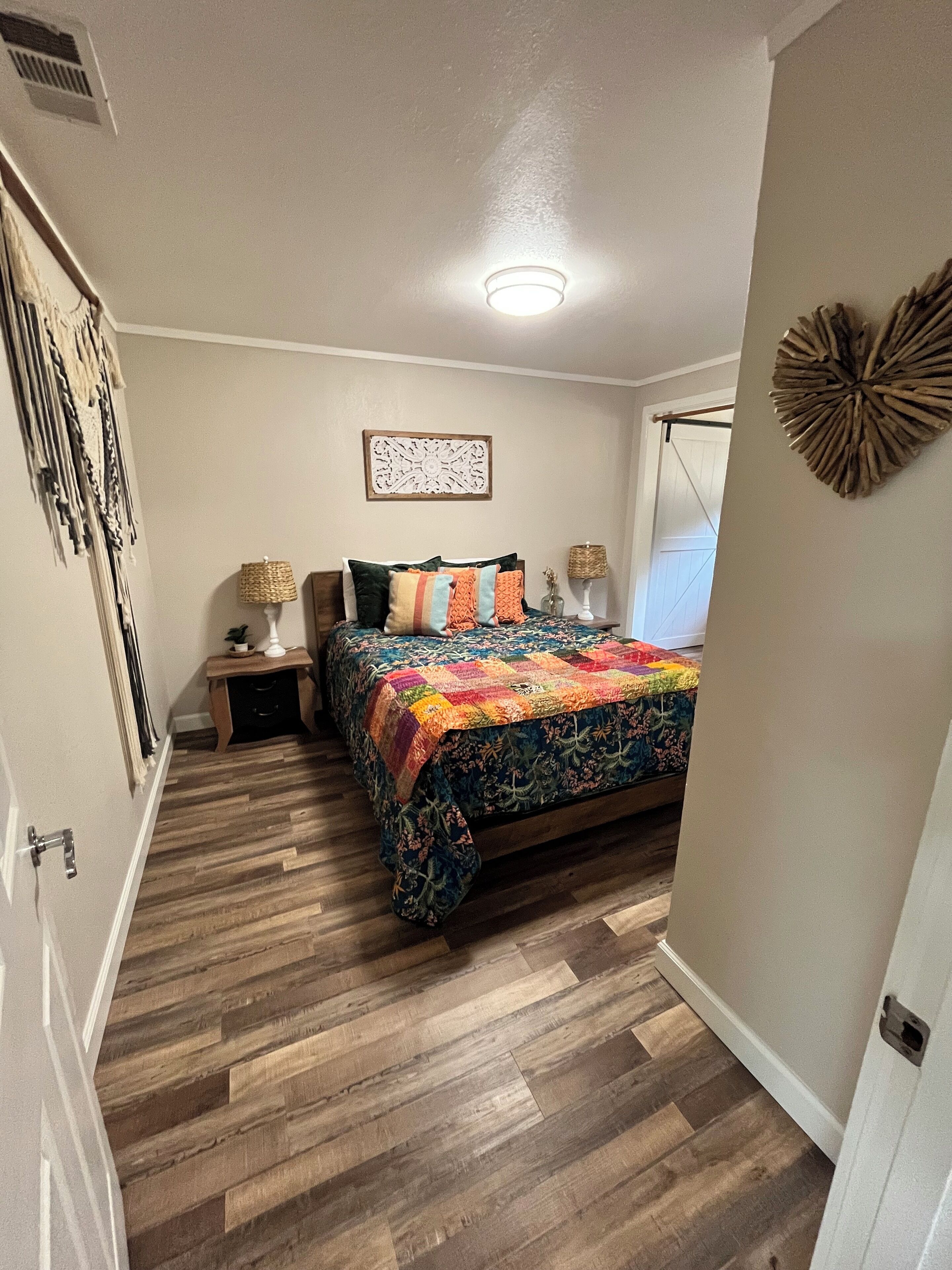 Un dormitorio con piso de madera, una cama con colcha colorida, dos mesitas de noche con lámparas, un adorno en la pared y una ventana con cortina.