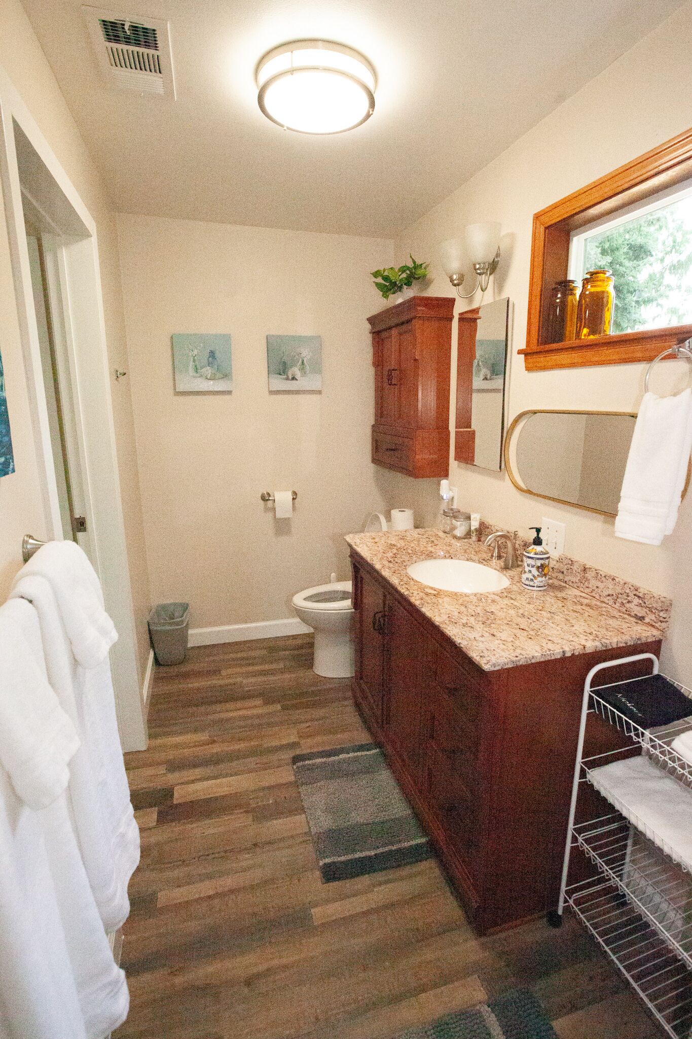Une salle de bain avec un meuble-lavabo en bois, un évier, des toilettes et un miroir. Deux tableaux encadrés sont accrochés au mur, ainsi qu’une poubelle et un porte-serviettes.