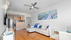 A modern living room with a white sofa, a glass coffee table, and a painting of a beach scene. - The Pool Resort - One Bedroom Pool & Ocean Views (Tweed)