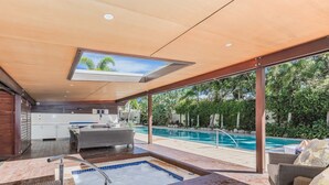 A modern outdoor pool area with a hot tub, lounge seating, and a kitchen area visible through large glass doors.
