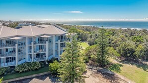 A modern multi-story building with large windows and balconies, surrounded by lush greenery and a view of the ocean.