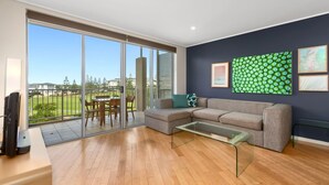 Ein modernes Wohnzimmer mit einer Glastür zum Balkon, ein graues Ecksofa, ein Glastisch und Blick auf eine Wohngegend.