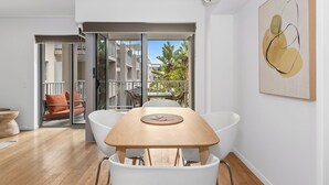 A modern interior with a wooden dining table, white chairs, and a large window with a view of a balcony and greenery. - The Sand Resort Kingscliff - One Bed Pool view (3202) (Tweed)