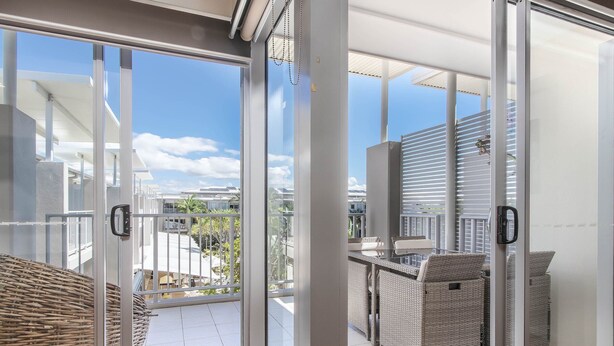 A balcony with wicker furniture and a glass door leading to an outdoor area with a view of other buildings and a clear sky. - The Sand Resort Kingscliff - One Bed Pool view (3302) (Tweed)