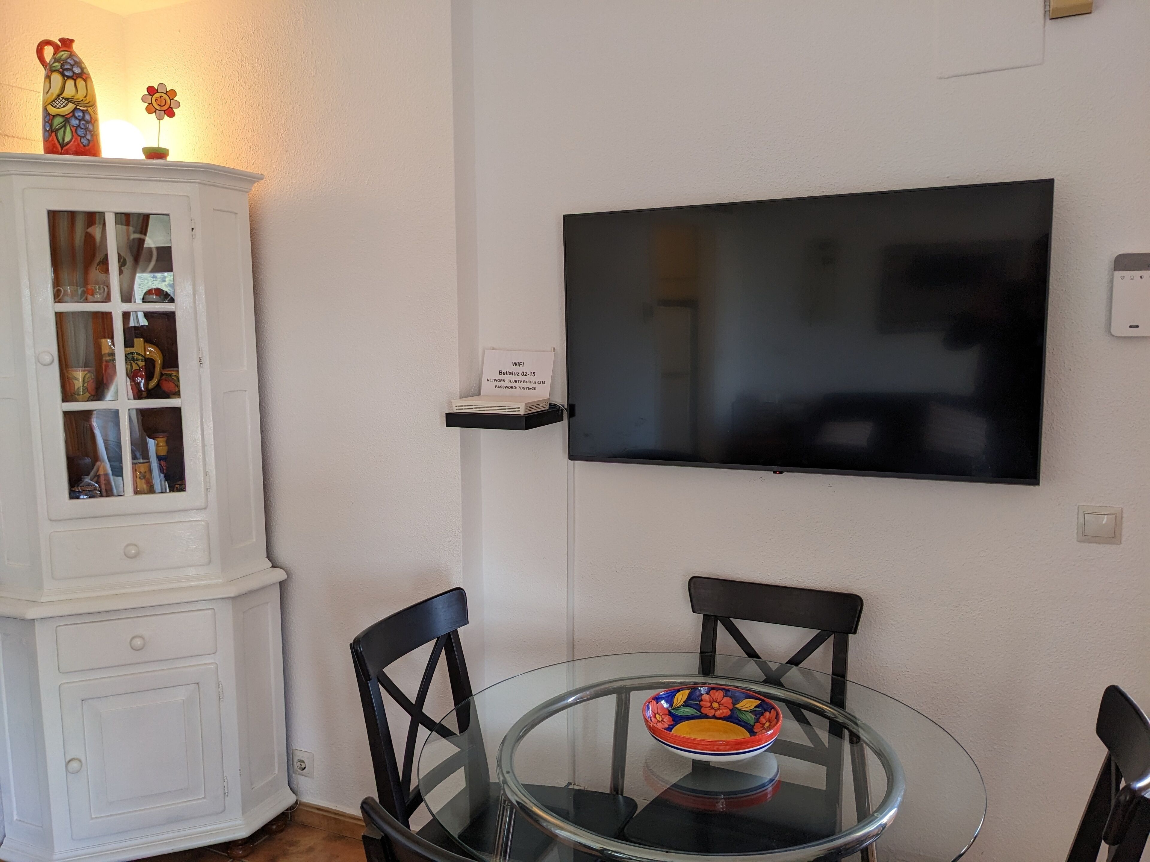 Un coin repas comprenant une table en verre, deux chaises, un téléviseur fixé au mur et un meuble contenant des objets décoratifs.
