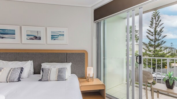 Un dormitorio moderno con una cama grande, una mesita de noche con lámpara y un ventanal corredizo que da a un balcón con vistas a árboles y edificios.