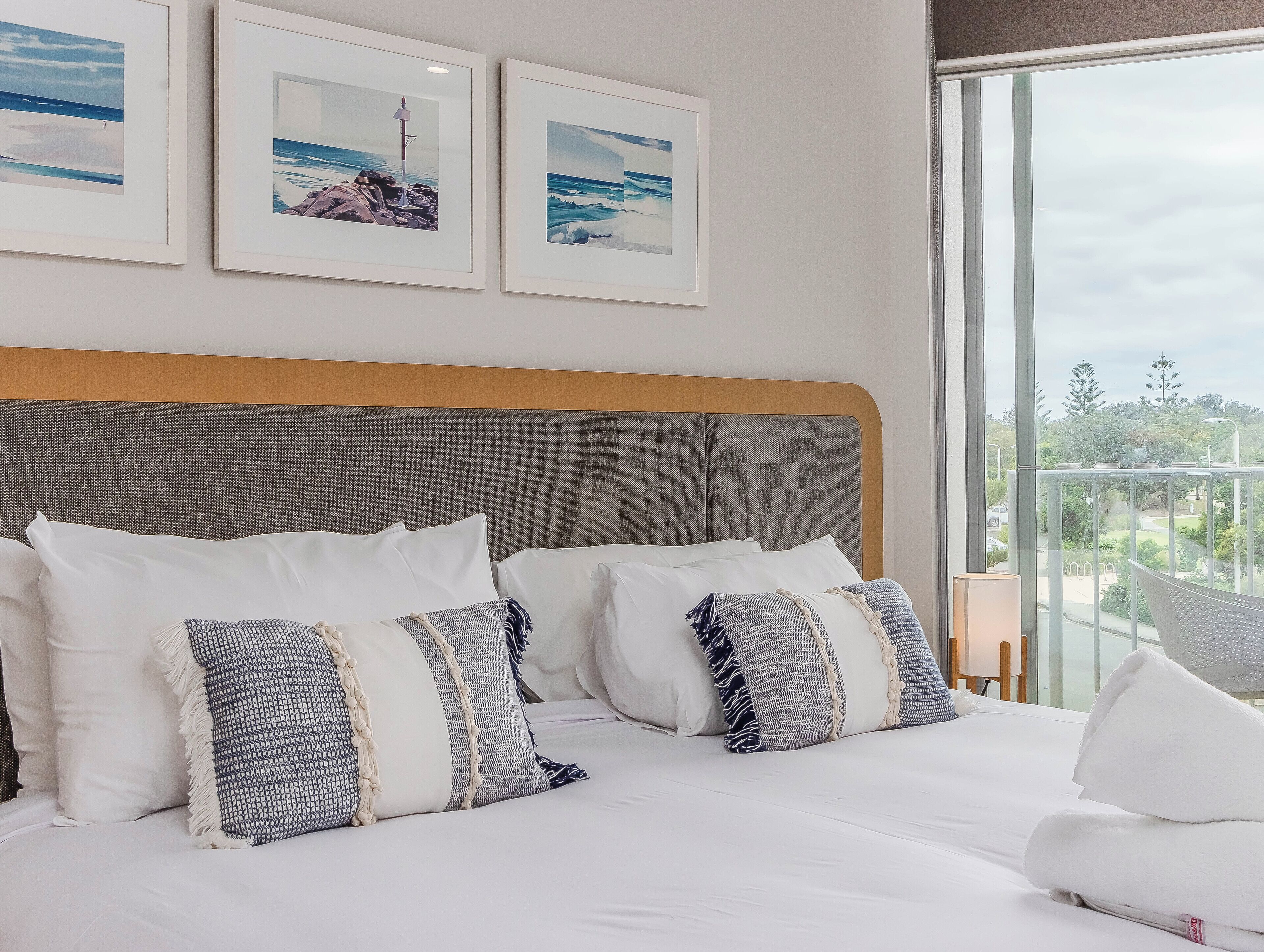 Una cama bien hecha con almohadas blancas y estampadas, cabecera gris y cuadros con motivos costeros en la pared.