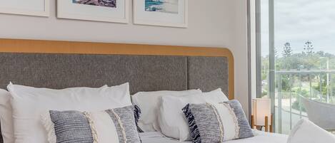 Una cama bien hecha con almohadas blancas y estampadas, cabecera gris y cuadros con motivos costeros en la pared.
