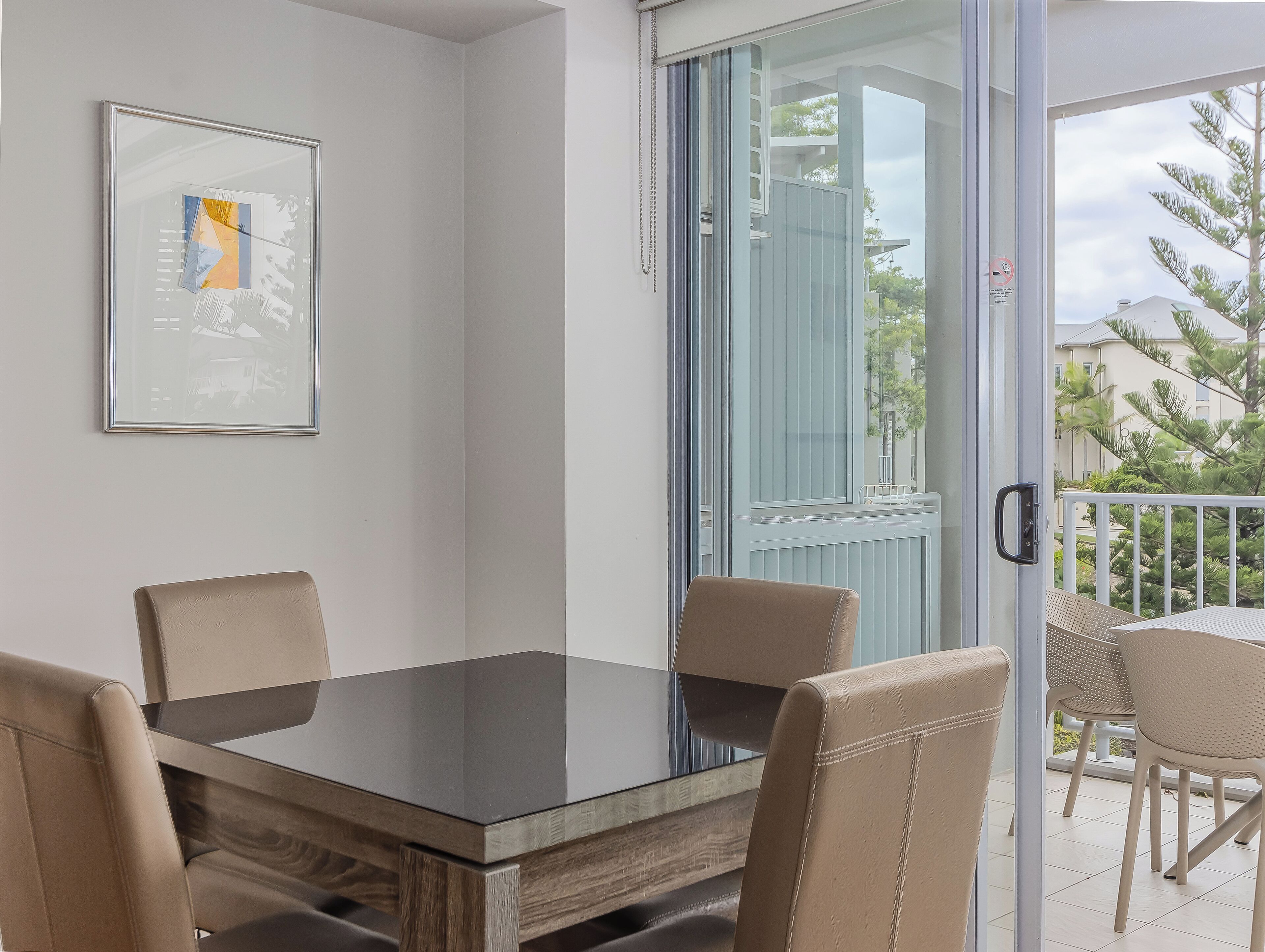 A dining area with a glass table and four chairs, a framed picture on the wall, and a view of a balcony with outdoor furniture.