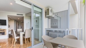A modern living room with a glass door leading to a dining area, a white air conditioning unit mounted on the wall, and a balcony with a metal railing. - The Sand Resort Kingscliff -One Bed Spa Suite (4202) (Tweed)