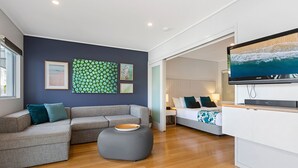 A modern living room with a grey sectional sofa, a grey ottoman, and a flat-screen TV mounted on a white cabinet. - The Sand Resort Kingscliff - Dual key 2B (6217/18) (Casuarina)