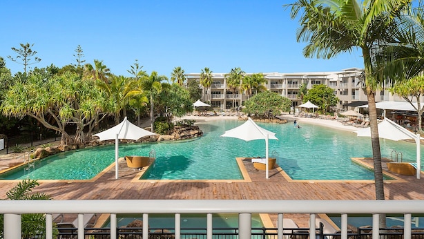 A resort pool area with multiple swimming pools, palm trees, and a building with balconies in the background. - The Sand Resort Kingscliff - Dual key 2B (6217/18) (Casuarina)